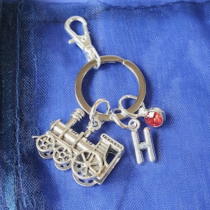 May include: Silver charm keychain featuring a miniature train engine, a red crystal charm, and a silver letter 'H' charm.