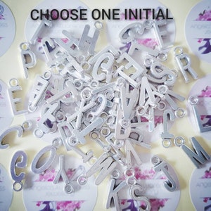 May include: A large pile of silver metal letter charms, each with a hole for attaching to a necklace or bracelet. The letters are all uppercase and are arranged randomly. The text "CHOOSE ONE INITIAL" is visible at the top of the image.