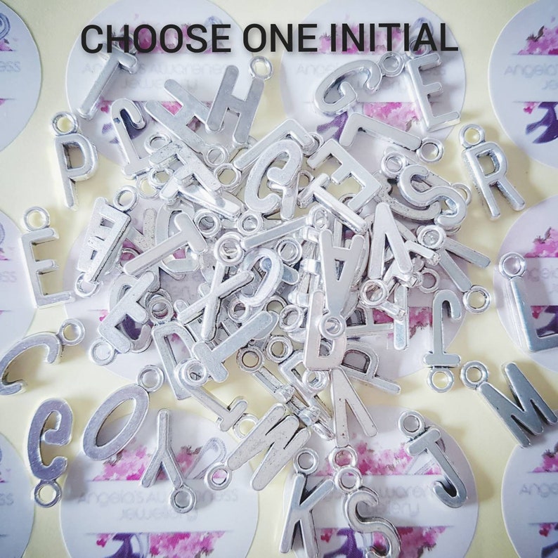 May include: A large pile of silver metal letter charms, each with a hole for attaching to a necklace or bracelet. The letters are all uppercase and are arranged randomly. The text "CHOOSE ONE INITIAL" is visible at the top of the image.