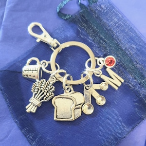 May include: Silver keyring with charms including a measuring cup, wheat stalks, a loaf of bread, a spoon, and a letter M with a red rhinestone.