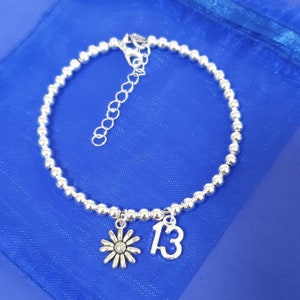 May include: A silver beaded bracelet with a silver daisy charm and a silver charm with the number 13.