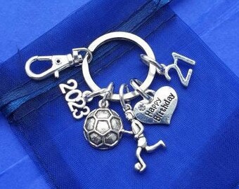 May include: Silver keychain with a soccer ball charm, a 'Happy Birthday' heart charm, a '21' charm, and a '2023' charm. The keychain is on a blue background.