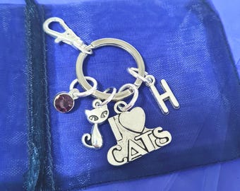 May include: Silver keychain with a charm that says "I love cats" and a small cat charm. The keychain also has a purple gem and a letter H charm. The keychain is on a blue background.