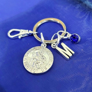 May include: Silver-coloured keychain featuring a Saint Christopher medal, an initial "M" charm, and a blue gemstone. The keychain includes a clasp and a keyring. The background is a solid blue fabric.