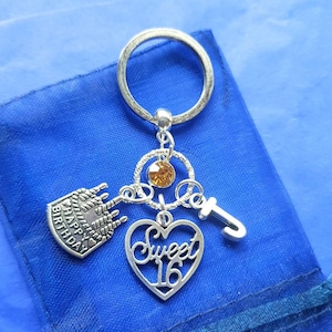 May include: Silver-coloured keychain with a 'Sweet 16' heart charm, a 'Happy Birthday' cake charm, and a 'J' charm. The keychain also features a gold-coloured gem and a circular key ring. The background is a blue fabric.