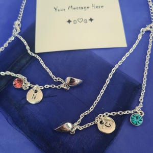 May include: Two silver necklaces with personalised pendants. One necklace has a red gemstone and a disc with the letter "N". The other has a teal gemstone and a disc with the letter "C". A card with the text "Your Message Here" is in the background.