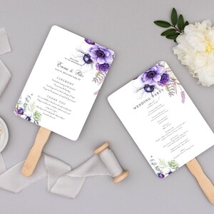 May include: Two white wedding fan programs with purple floral accents and wooden handles. One fan reads "Emma & Phil" and the other reads "Wedding Party". A white flower, ribbon, and wax seal are also present.