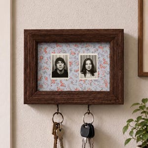 May include: A rectangular dark brown wooden wall-mounted frame featuring a light blue background with a small red floral pattern. Two black and white portrait photographs are displayed inside. Two metal hooks at the bottom hold two sets of keys.