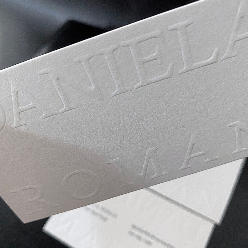May include: Close-up of white business cards with embossed text. The name "DANIELA ROMAN" is prominently displayed in a serif font. The cards have a textured surface and are stacked, with a dark background.
