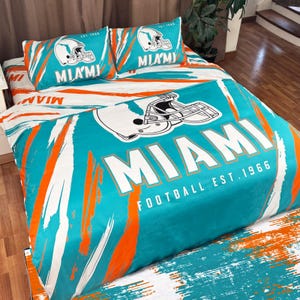 Könnte beinhalten: Ein Bettwäsche-Set im Miami Dolphins-Design in Türkis, Orange und Weiß. Das Set enthält einen Bettbezug und zwei Kissenbezüge, alle mit dem Logo des Teams, einem Helm und dem Wort "MIAMI". Der Text "FOOTBALL EST. 1966" ist ebenfalls sichtbar.