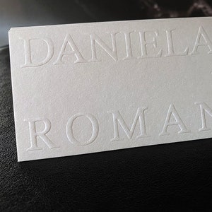 May include: A stack of white business cards with the embossed text "DANIELA ROMAN" in a serif font. The cards are on a black surface, with the top card in focus.