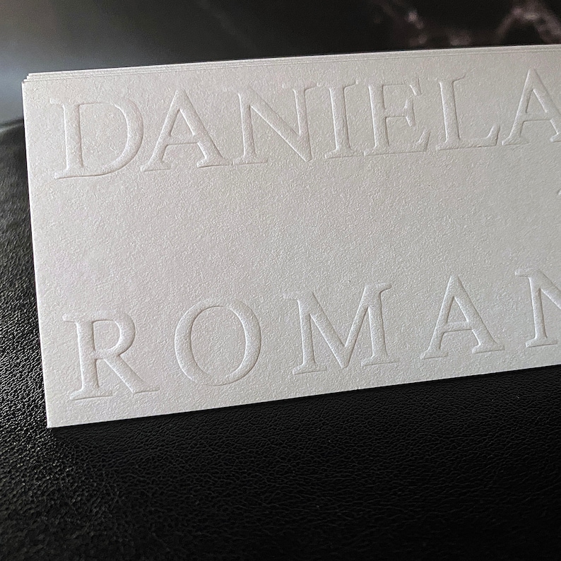 May include: A stack of white business cards with the embossed text "DANIELA ROMAN" in a serif font. The cards are on a black surface, with the top card in focus.