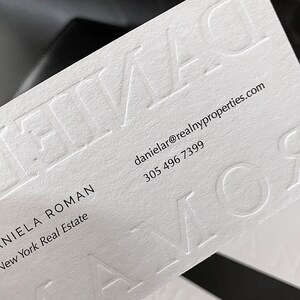 May include: A white business card with embossed lettering. The card displays the name "DANIELA ROMAN" and contact details, including an email address and a phone number. The business is "New York Real Estate".