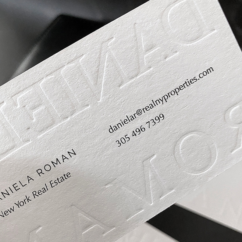May include: A white business card with embossed lettering. The card displays the name "DANIELA ROMAN" and contact details, including an email address and a phone number. The business is "New York Real Estate".