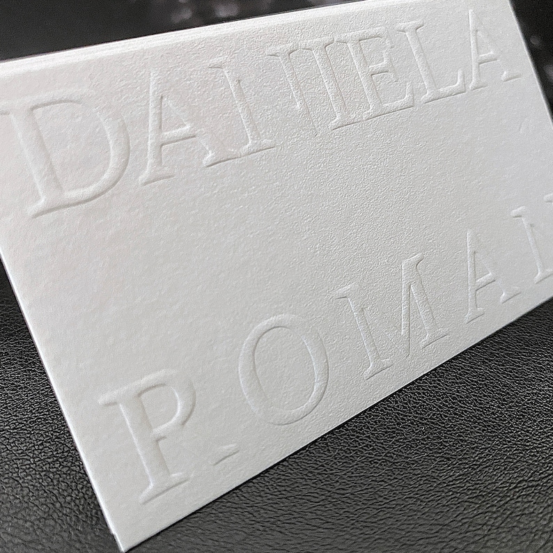 May include: A white business card featuring the embossed name "DANIELA ROMAI". The card has a textured surface and is angled, showing the raised lettering. The background is a dark surface, possibly leather.