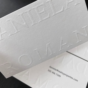 May include: Close-up of white business cards with embossed text. The cards feature the name "DANIELA ROMAN" in large, bold letters. Contact information, including an email address and phone number, is printed in a smaller font.