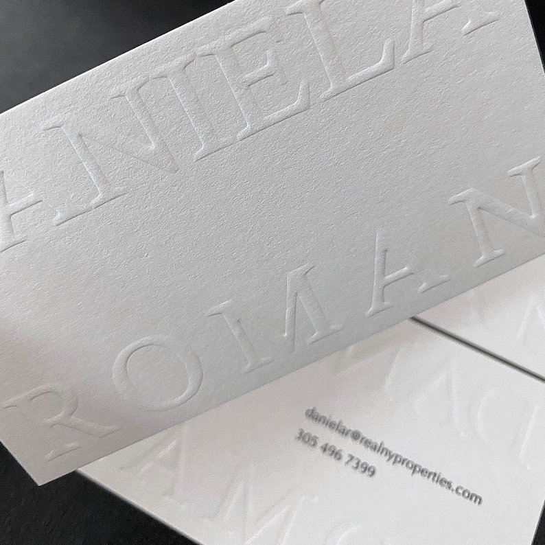 May include: Close-up of white business cards with embossed text. The cards feature the name "DANIELA ROMAN" in large, bold letters. Contact information, including an email address and phone number, is printed in a smaller font.