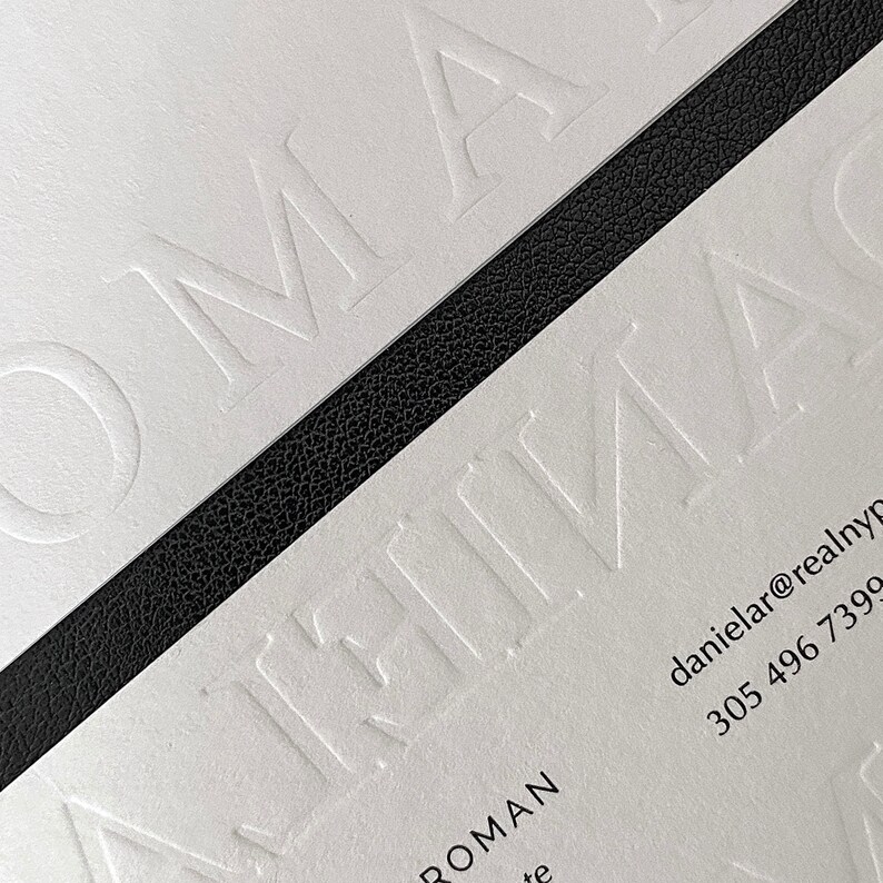 May include: Close-up of white business cards with embossed text. A black textured strip bisects the cards. The text includes the name "ROMAN" and contact details. The design is minimalist and modern.