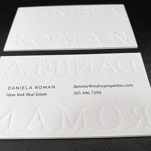 May include: Two white business cards with embossed text. The top card displays the name "DANIELA ROMAN" in large, raised letters. The bottom card has the name and contact information, including an email address and phone number.
