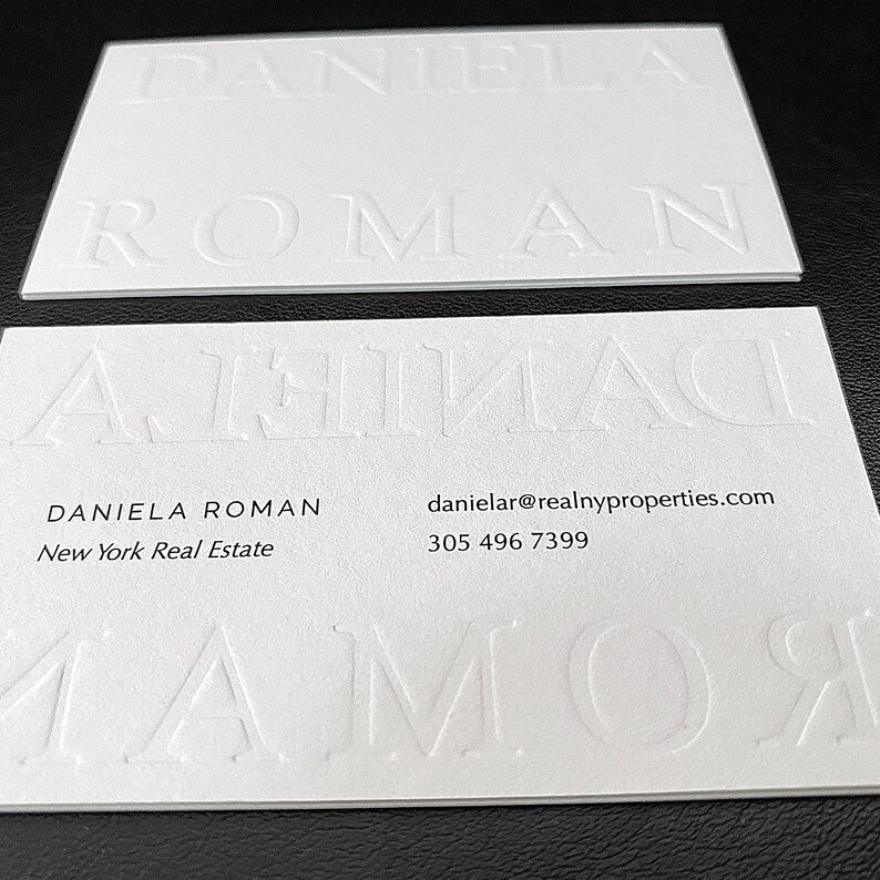 May include: Two white business cards with embossed text. The top card displays the name "DANIELA ROMAN" in large, raised letters. The bottom card has the name and contact information, including an email address and phone number.