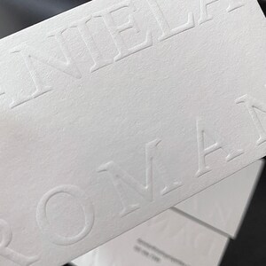 May include: Close-up of white business cards with embossed text. The cards are stacked, with the top card displaying the name "DANIEL ROMAN" in a sans-serif font. The paper has a textured finish.