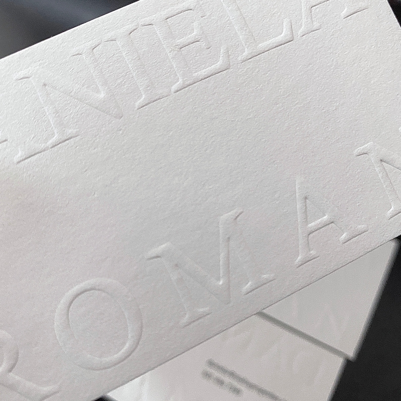 May include: Close-up of white business cards with embossed text. The cards are stacked, with the top card displaying the name "DANIEL ROMAN" in a sans-serif font. The paper has a textured finish.