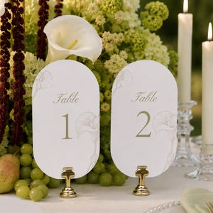 以下が含まれることがあります： Table 1とTable 2と書かれた2枚の白い楕円形のテーブルナンバーカード。薄茶色のカラーリリーの線画が描かれています。各カードは、磨かれた金色の小さな金属製スタンドで直立しており、緑色のブドウが置かれた表面に配置されています。