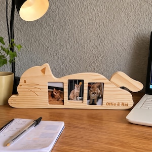 Könnte beinhalten: Ein heller Holzbilderrahmen in Form einer liegenden Katze mit drei rechteckigen Ausschnitten für Fotos. Der Rahmen zeigt eingravierte Schnurrhaare, ein spitzes Ohr, einen Schwanz und den Text Ollie & Kai auf der unteren rechten Seite.