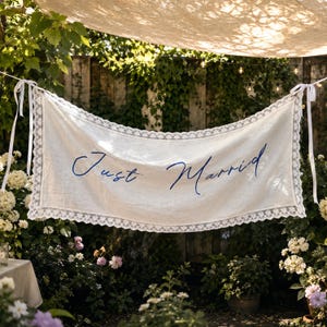 May include: A rectangular off-white fabric banner featuring a delicate white lace border and white ribbon ties at the corners. The text "Just Married" is embroidered in a dark blue cursive script across the center of the banner.