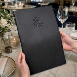 May include: A black rectangular menu cover featuring a debossed logo with a chef hat icon and the text Kitchen CAFE & RESTO. The cover has a smooth matte finish and is held upright, displaying the centered branding on the front surface.