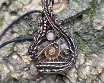 Puede incluir: Colgante artesanal con un amonita fosilizada, encapsulada en alambre de cobre. El colgante incluye una pequeña piedra de ópalo iridiscente. El trabajo de alambre es intrincado, con una mezcla de metales oscuros y cobrizos. El colgante cuelga de un cordón negro.