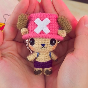 Puede incluir: Una figura de ganchillo hecha a mano de un personaje de dibujos animados, con un sombrero rosa y una cruz blanca, cuernos marrones y un cuerpo beige. Lleva pantalones cortos morados y tirantes azules. La figura se sostiene en manos abiertas.