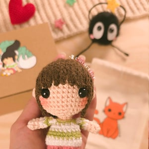 Puede incluir: Una muñeca amigurumi de crochet de una niña pequeña con cabello castaño, que lleva una camisa a rayas verdes y blancas y pantalones cortos rosas. La muñeca sostiene un pequeño gato de crochet en sus brazos.