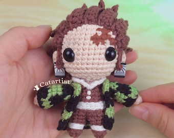 May include: Crocheted doll of a character with brown hair, a tan face, and large black eyes. The doll wears a brown and green outfit with black accents. The doll is held in two hands. A red crocheted heart is in the background.