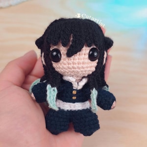 Puede incluir: Muñeca de ganchillo hecha a mano con cabello negro y ojos negros grandes. La muñeca viste un atuendo azul oscuro con detalles blancos y un chal verde claro. El texto "Catartist" es visible en la parte superior derecha.