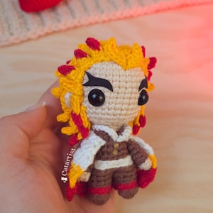 Puede incluir: Muñeca de ganchillo hecha a mano con cabello amarillo, rojo y beige, vestida con un atuendo marrón y blanco. La muñeca tiene ojos negros grandes y se sostiene en una mano. Un corazón rojo de ganchillo está en el fondo.