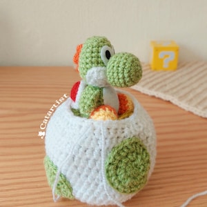 Può includere: Un giocattolo di Yoshi fatto a mano all'uncinetto, verde con cresta arancione e sella rossa, che esce da un uovo bianco con macchie verdi. Un blocco giallo con un punto interrogativo è sullo sfondo. Il giocattolo è su una superficie di legno.