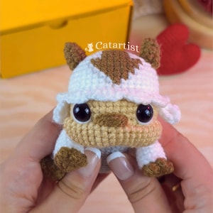 Peut inclure: Un petit jouet en peluche crocheté à la main avec un visage beige, de grands yeux noirs et une tête blanche ornée d'une marque en forme de flèche brune. La créature a des membres blancs avec des pattes et des oreilles brunes, texte Catartist.