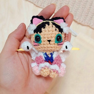Puede incluir: Un juguete de gato de crochet con orejas rosas, una capa con capucha blanca y detalles azules y rosas. El gato tiene el pelo negro y los ojos verdes.