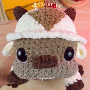 Puede incluir: Un peluche de ganchillo hecho a mano con un esquema de color crema y marrón con un patrón de flecha distintivo en la cabeza. La criatura tiene grandes ojos negros brillantes, una nariz marrón y orejas pequeñas. El texto Catartist es visible.
