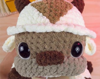 May include: A handmade crochet plush toy featuring a cream and brown color scheme with a distinct arrow pattern on the head. The creature has large, glossy black eyes, a brown nose, and small brown ears. The text Catartist is visible above the toy.