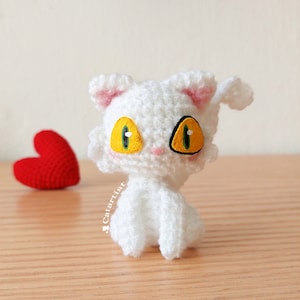 Puede incluir: Gato blanco de crochet con ojos amarillos y pupilas verdes. El gato está sentado en una superficie de madera con un corazón rojo de crochet en el fondo.