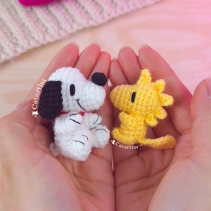 May include: Two small, handmade crochet figures of Snoopy and Woodstock are held in cupped hands. Snoopy is white with black ears and a black spot, wearing a red collar. Woodstock is yellow. The background is a soft pink and white.
