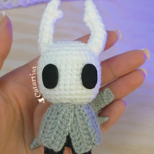 Puede incluir: Una figura de ganchillo hecha a mano de un personaje con cabeza blanca, ojos negros grandes y dos cuernos blancos. La figura lleva una capa gris y tiene pequeños pies negros. El texto "Catartist" es visible.