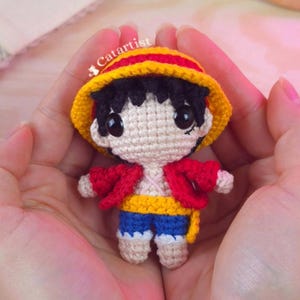 Puede incluir: Muñeca de ganchillo hecha a mano de un personaje con sombrero amarillo y rojo, chaqueta roja, pantalones cortos azules y cinturón amarillo. La muñeca tiene pelo negro y ojos grandes y negros. El texto "Catartist" es visible.