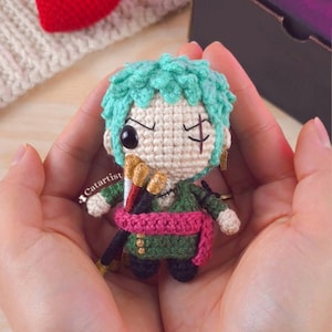 Puede incluir: Una figura de ganchillo hecha a mano de un personaje con cabello verde, un abrigo verde y una faja rosa. La figura sostiene tres espadas y tiene una cicatriz debajo de un ojo. La figura se sostiene con dos manos. El nombre del artista, "Catartist", es visible.