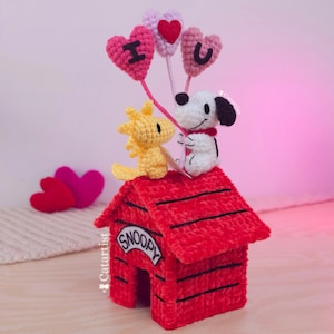 Puede incluir: Una caseta para perros Snoopy hecha a mano con ganchillo con figuras de Snoopy y Woodstock. Snoopy sostiene globos en forma de corazón que deletrean "I U". La caseta roja para perros tiene la palabra "Snoopy". Las figuras están sobre un fondo rosa claro.