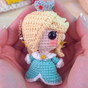 Puede incluir: Muñeca de ganchillo hecha a mano de un personaje con cabello rubio, un vestido azul y una corona. La muñeca tiene ojos de cuentas negras y mejillas rosadas. La muñeca se sostiene en manos abiertas. Es un personaje de videojuego.