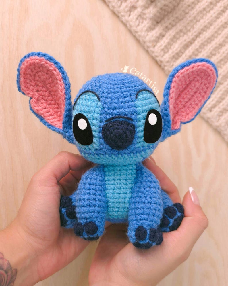 K&ouml;nnte beinhalten: Blau-rosa geh&auml;kelte Stitch-Puppe mit gro&szlig;en schwarzen Augen und Ohren. Die handgefertigte Puppe hat ein strukturiertes Aussehen. Die Puppe ist eine Figur aus dem Disney-Film Lilo & Stitch.