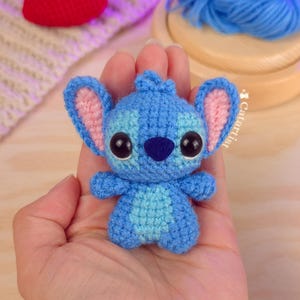 Könnte beinhalten: Eine handgefertigte blaue Stitch-Puppe aus Häkelarbeit mit großen schwarzen Augen und rosa Innenohren. Das kleine Plüschtier wird in einer Hand gehalten, was seine detaillierte Handwerkskunst zeigt. Im Hintergrund ist ein rotes Häkelherz zu sehen.